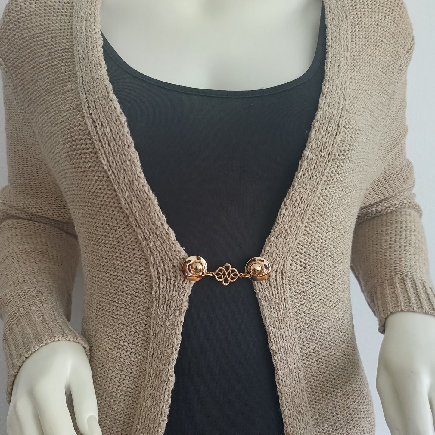 Goudkleurige vestsluiting met sierlijk verbindingsmotief, handgemaakt en zacht van uitstraling, bevestigd aan een beige vest.