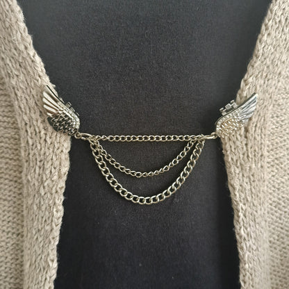 Close-up van een zilverkleurige vestsluiting met verfijnde vleugels en drie elegant vallende kettingen, die vrijheid en balans uitstralen.