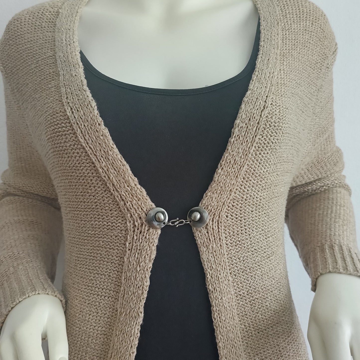 “Zilverkleurige vestsluiting De Twist met sierlijk haak-en-oog ontwerp, gedragen op een beige vest voor een elegante en subtiele uitstraling.”