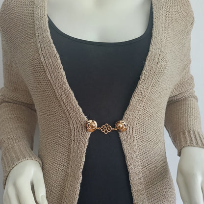 Goudkleurige vestsluiting met sierlijk verbindingsmotief, handgemaakt en zacht van uitstraling, bevestigd aan een beige vest.
