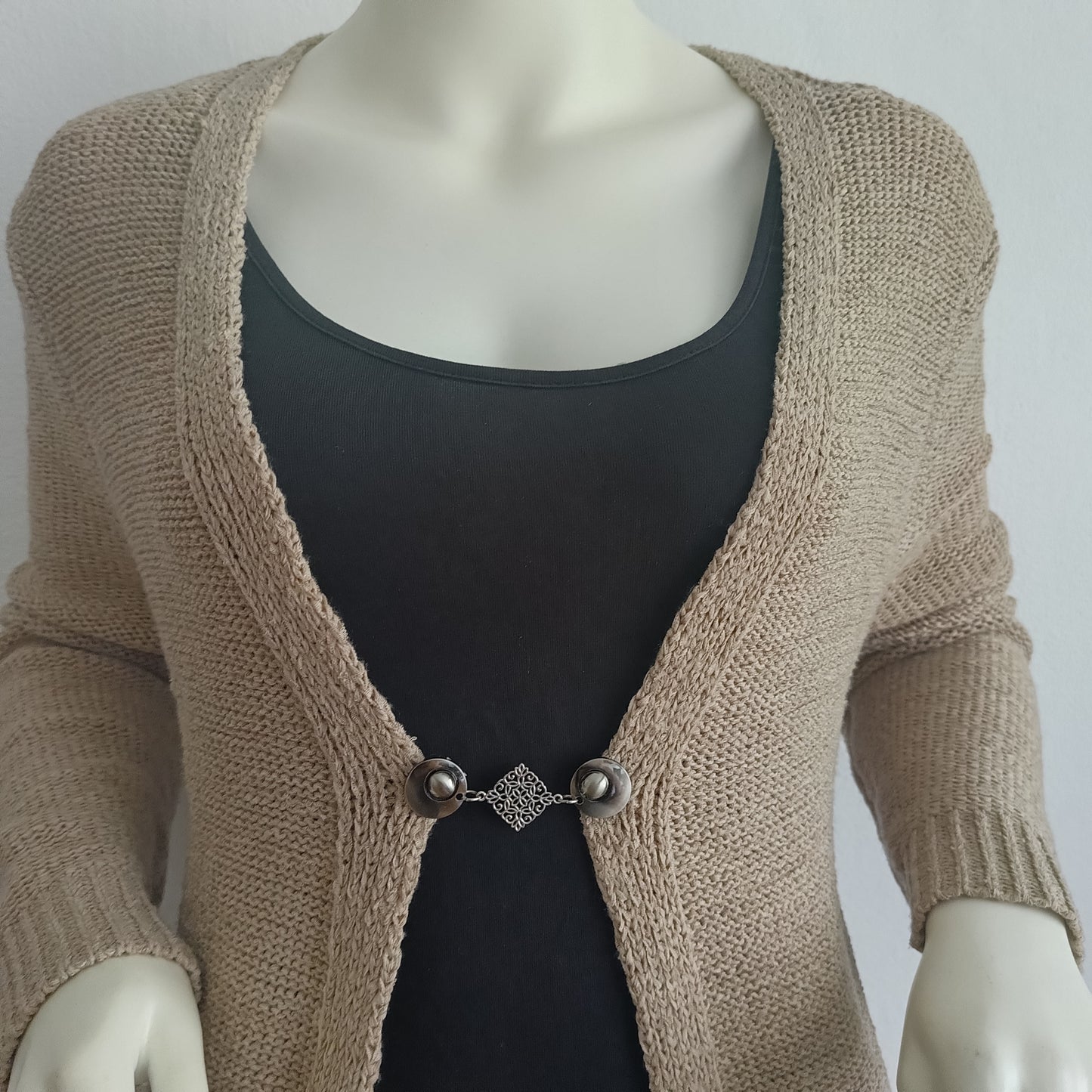 Zilverkleurige Fashionclip vestsluiting met sierlijk decoratief middenstuk, bevestigd op een beige vest voor een elegante en subtiele uitstraling.
