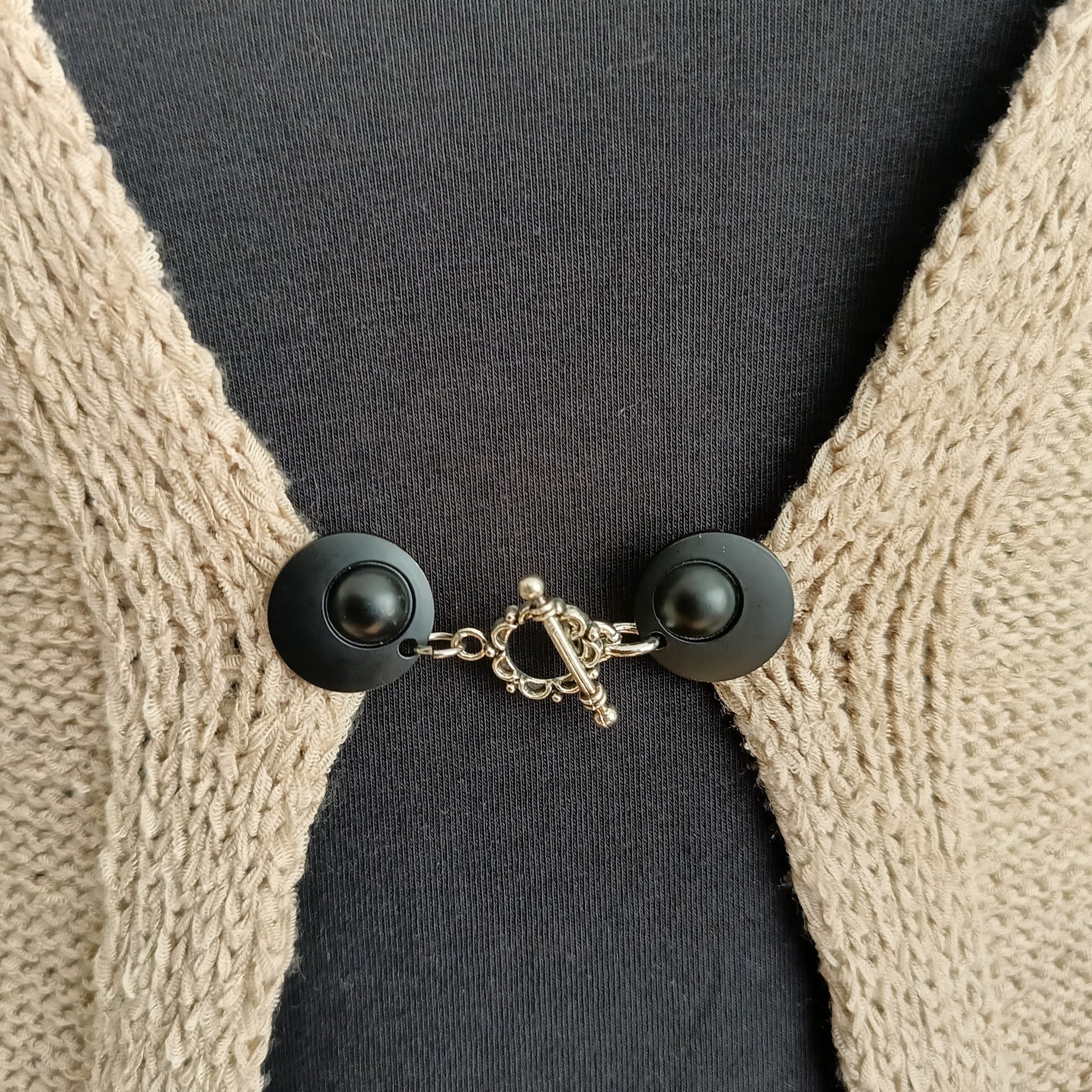 Vestsluiting met mat zwarte clips en een zilverkleurige bloemrand-bedel, handgemaakt voor een elegante en moderne uitstraling.