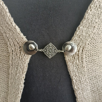 Zilverkleurige vestsluiting Decoratie met filigraan diamantvorm, bevestigd aan een beige vest.