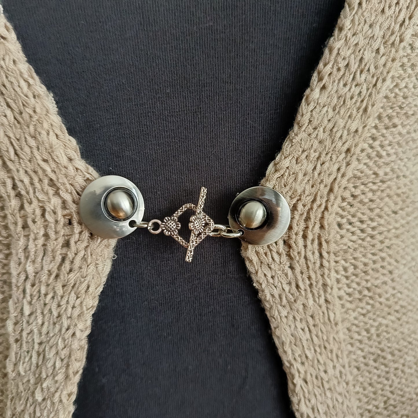 Een elegante zilverkleurige vestclip met een opengewerkte ruitvorm, bevestigd aan twee ronde magneten. De clip sluit het vest subtiel en geeft uw outfit een verfijnde uitstraling.