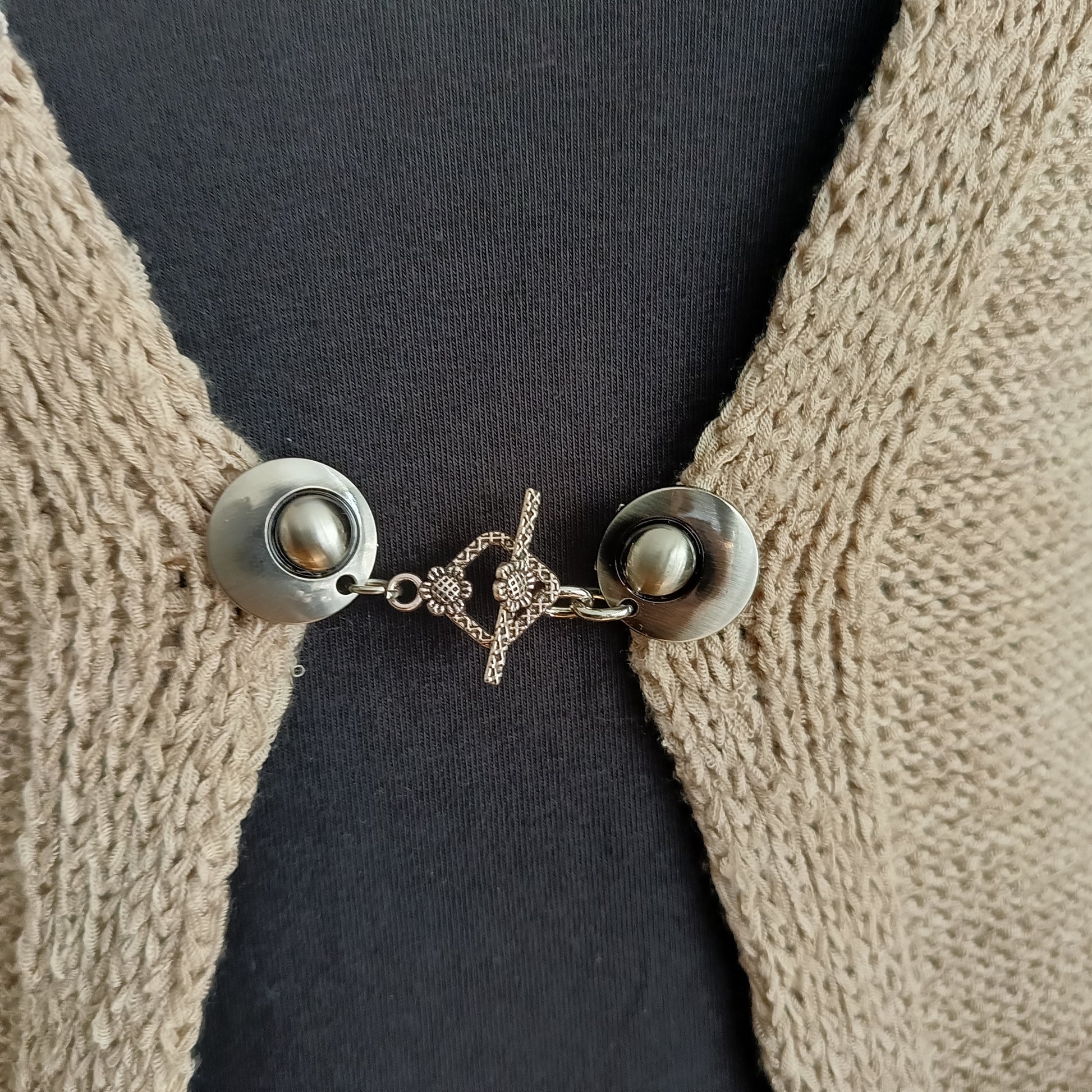 Een elegante zilverkleurige vestclip met een opengewerkte ruitvorm, bevestigd aan twee ronde magneten. De clip sluit het vest subtiel en geeft uw outfit een verfijnde uitstraling.