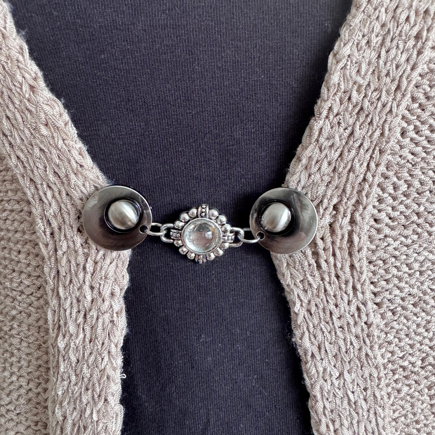Vestsluiting De Kameleon in close-up op een gebreid vest, elegante Fashionclip met zilverkleurige clips en glinsterende siersteen tussenbedel.
