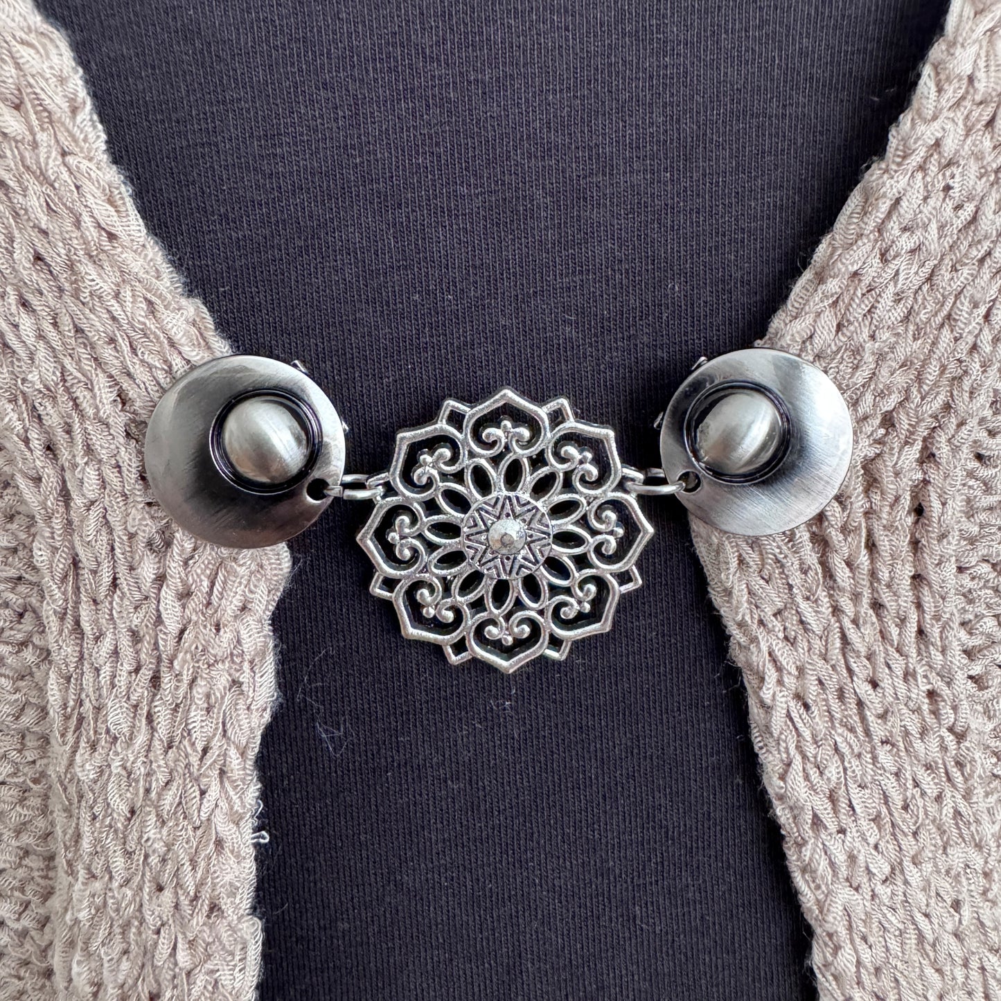 Vestsluiting De Mandala in close-up op een gebreid vest, elegante Fashionclip met zilverkleurige clips en opengewerkte mandala tussenbedel.