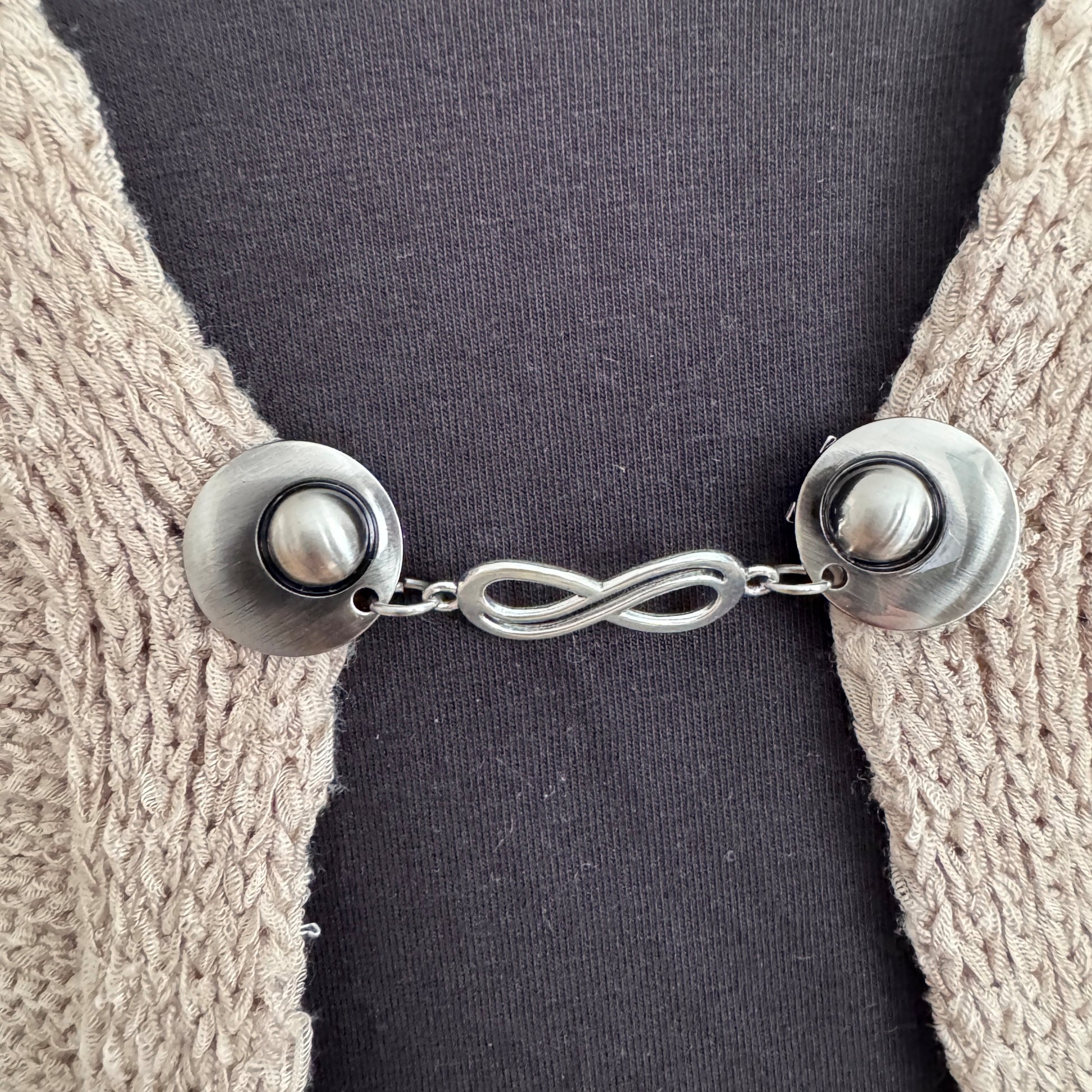 Vestsluiting De Oneindigheid in close-up op een gebreid vest, elegante Fashionclip met zilverkleurige clips en infinity tussenbedel.