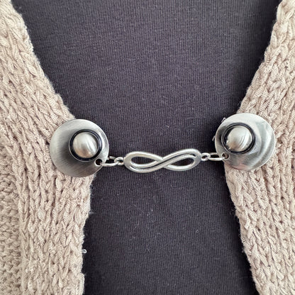 Vestsluiting De Oneindigheid in close-up op een gebreid vest, elegante Fashionclip met zilverkleurige clips en infinity tussenbedel.