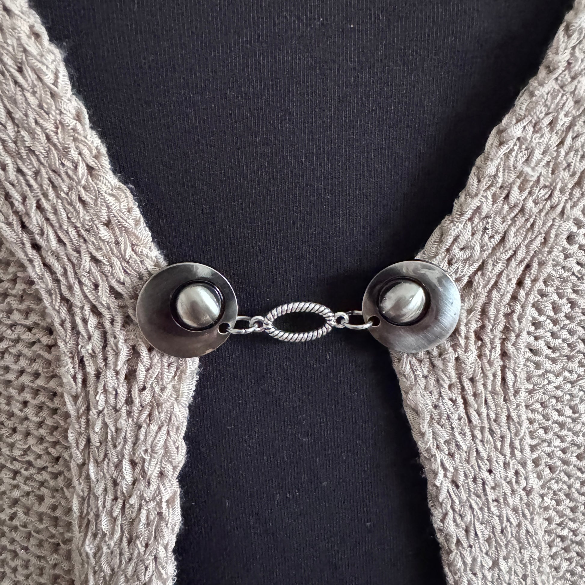 Vestsluiting De Ovaal in close-up op een gebreid vest, elegante Fashionclip met zilverkleurige clips en ovale tussenbedel.