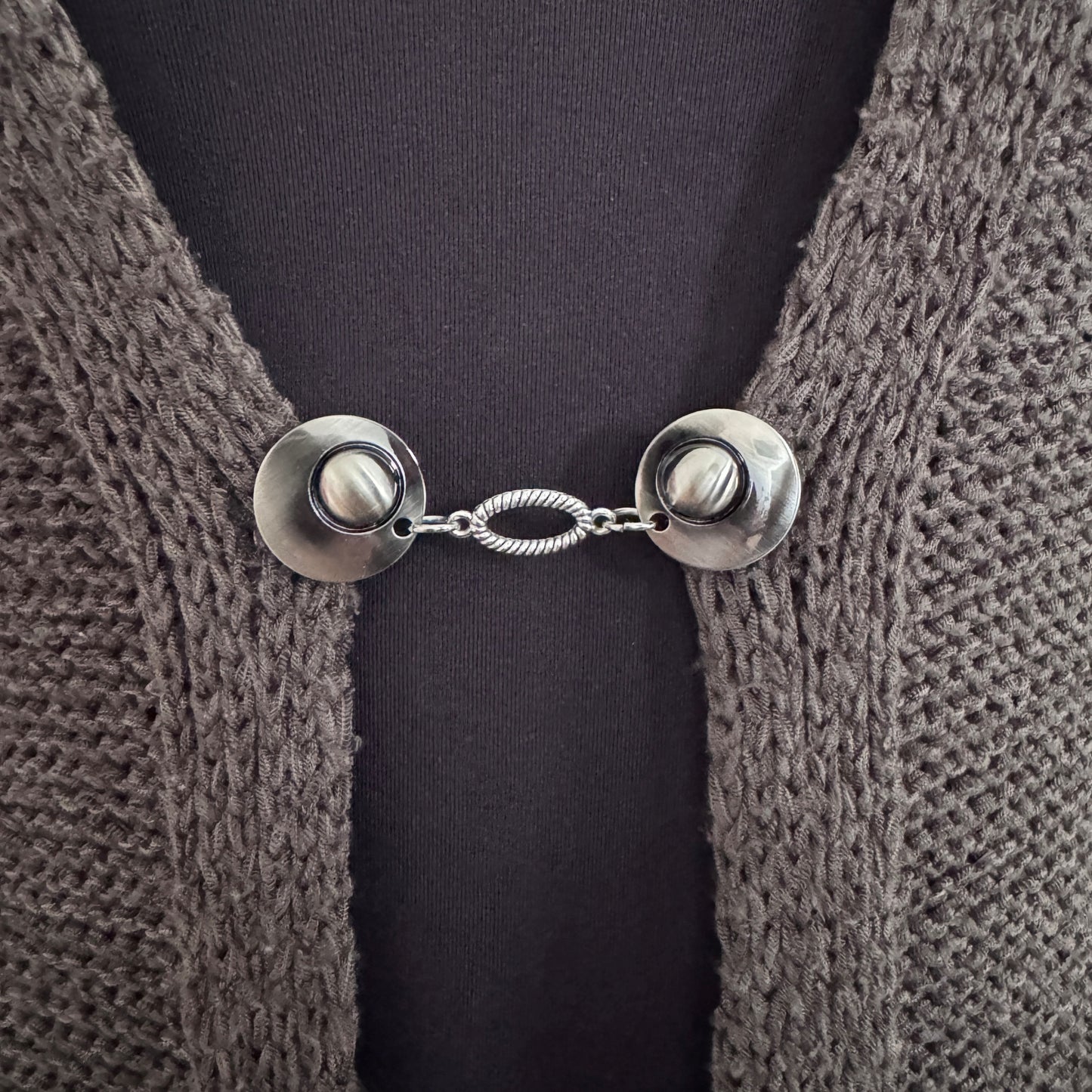 Vestsluiting De Ovaal in close-up op een gebreid vest, elegante Fashionclip met zilverkleurige clips en ovale tussenbedel.