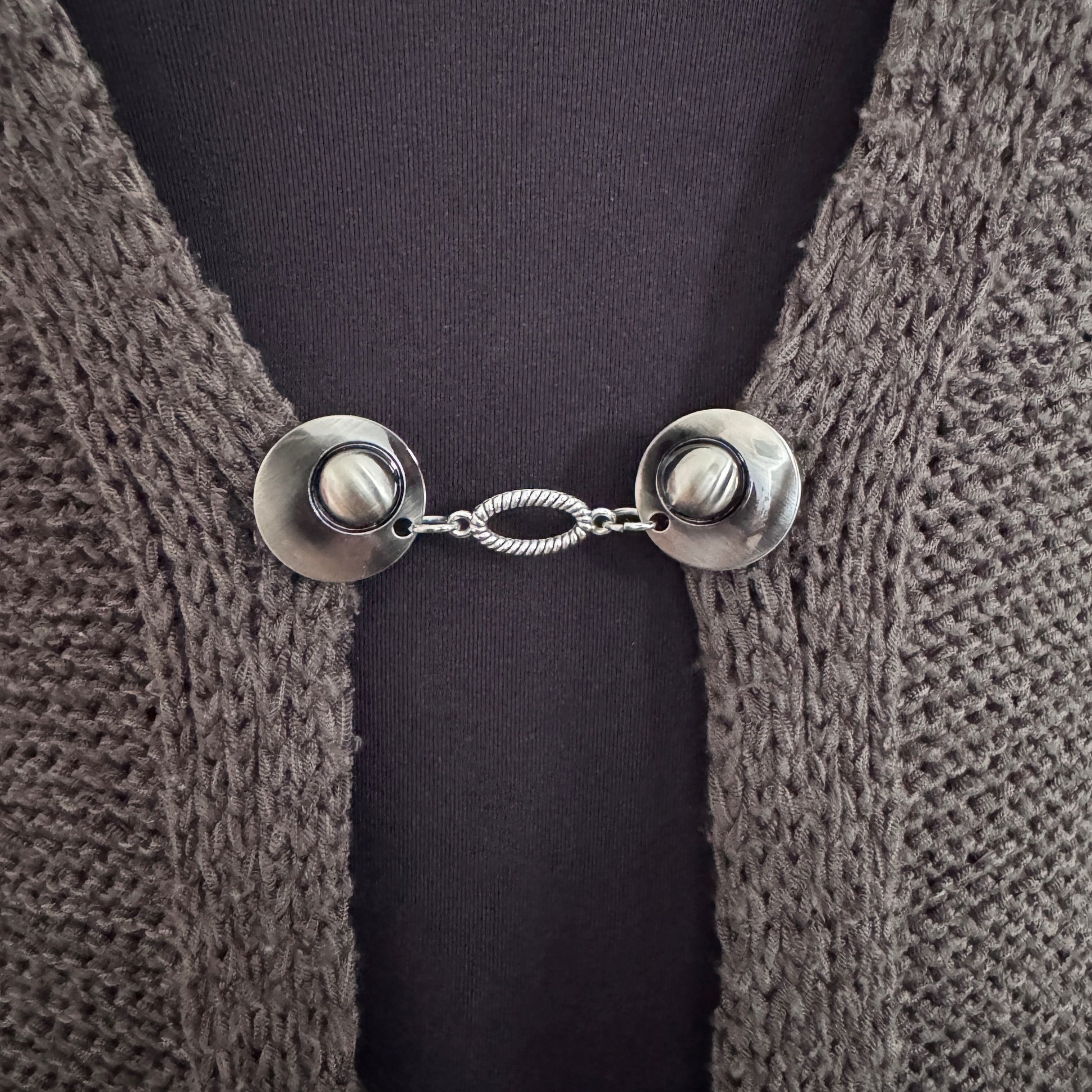 Vestsluiting De Ovaal in close-up op een gebreid vest, elegante Fashionclip met zilverkleurige clips en ovale tussenbedel.