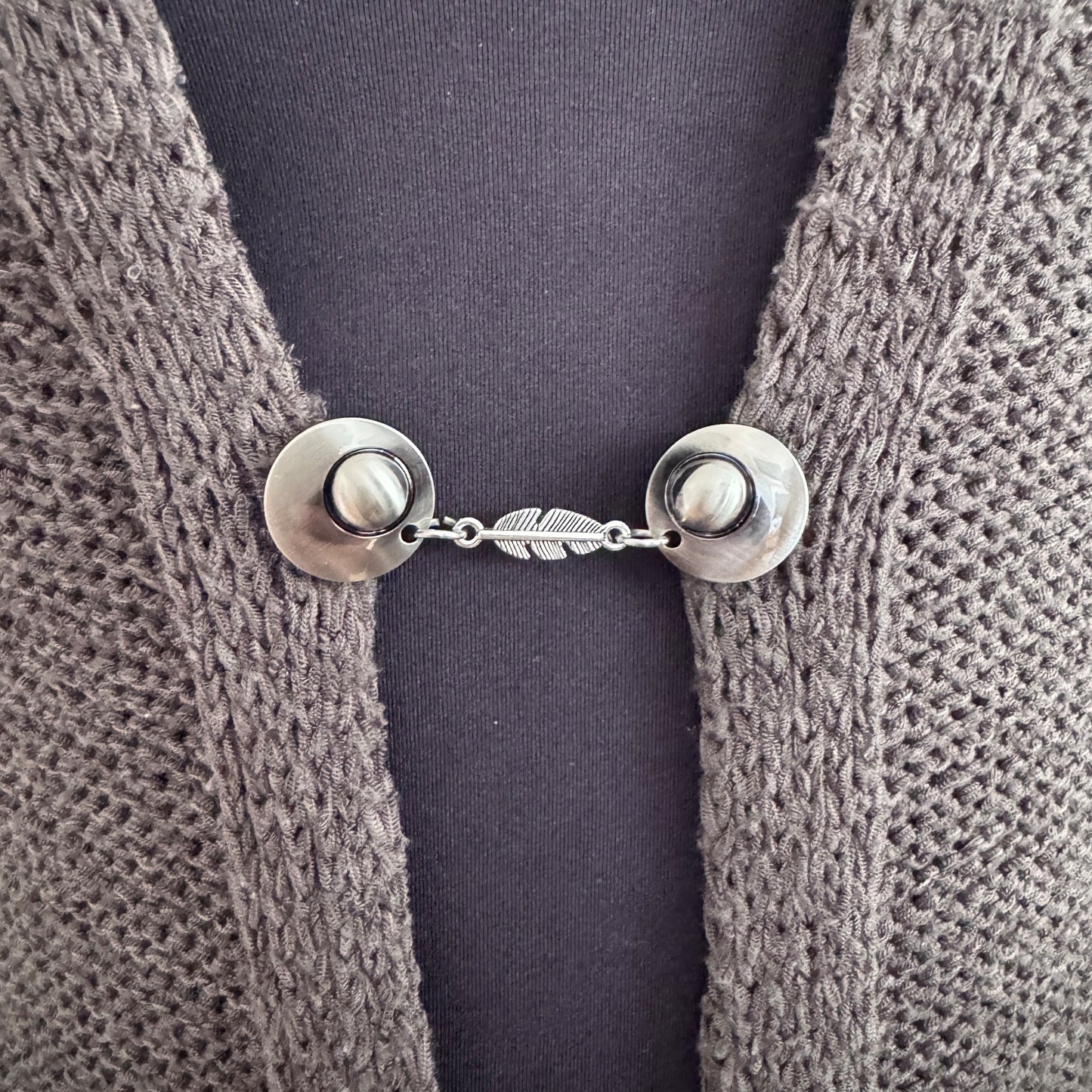 Vestsluiting De Veer in close-up op een gebreid vest, elegante Fashionclip met zilverkleurige clips en verfijnde veer tussenbedel.