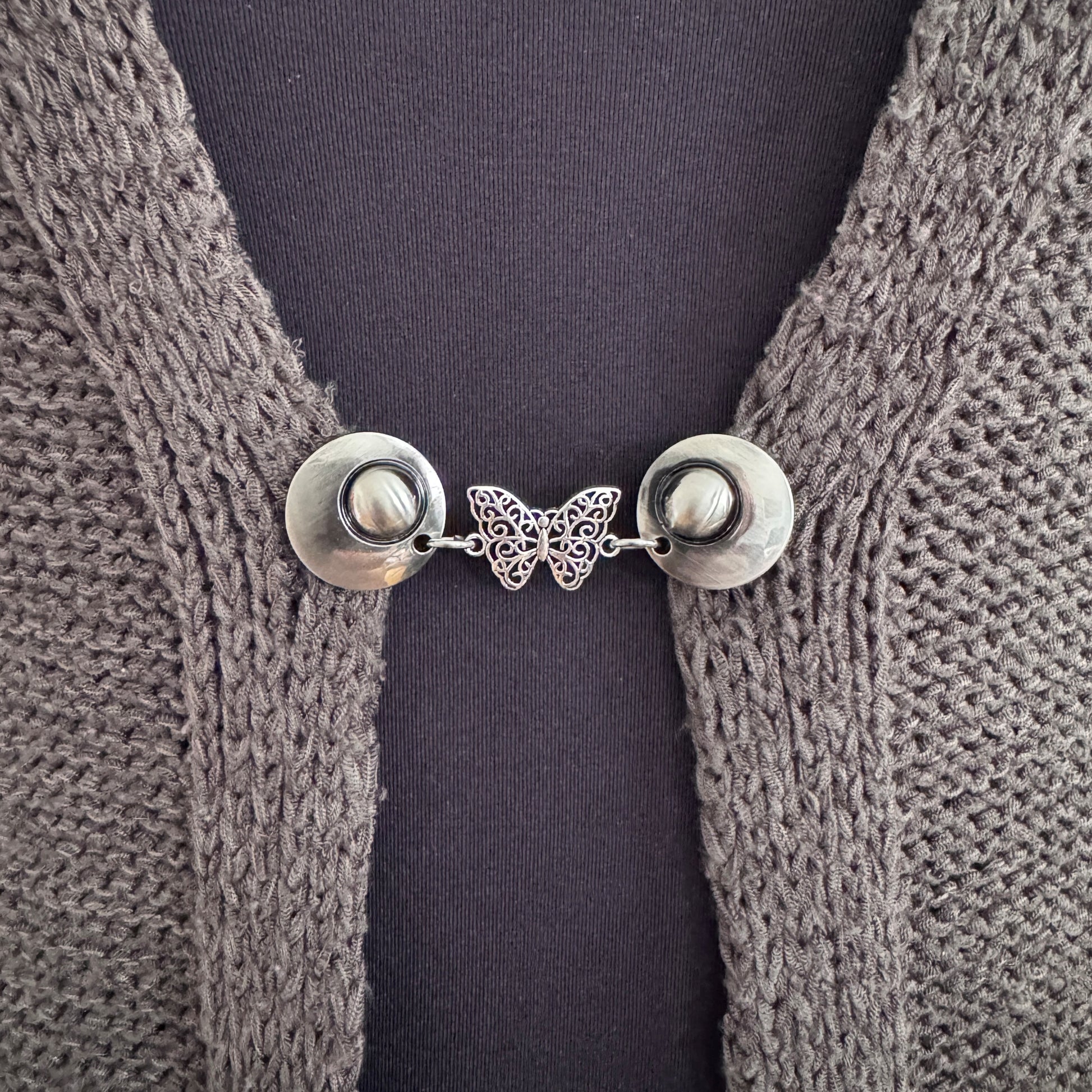 Vestsluiting De Vlinder in close-up op een gebreid vest, elegante Fashionclip met zilverkleurige clips en opengewerkte vlinder tussenbedel.