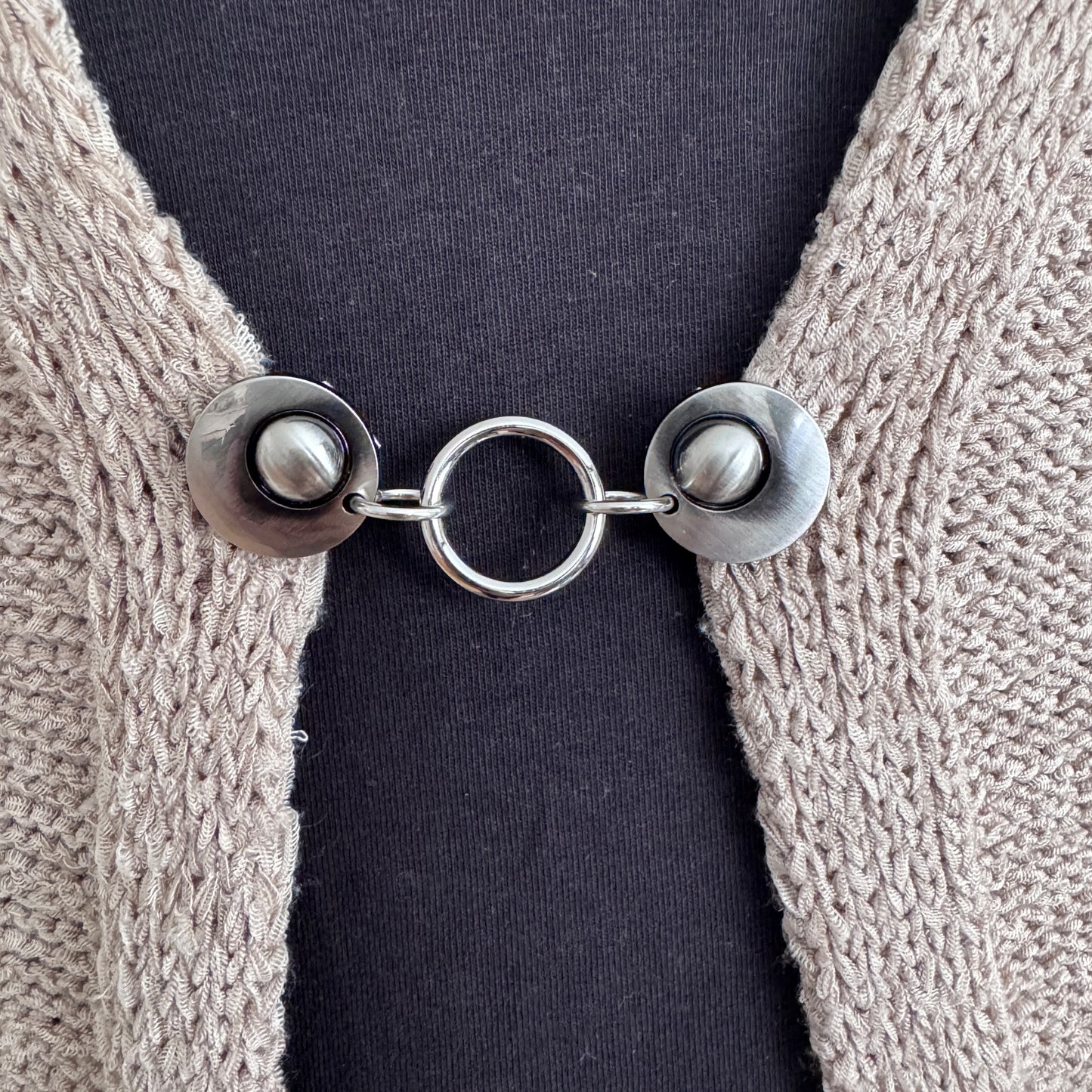 Vestsluiting Het Rondje zilver in close-up op een gebreid vest, elegante Fashionclip met zilverkleurige clips en ronde ringverbinding.