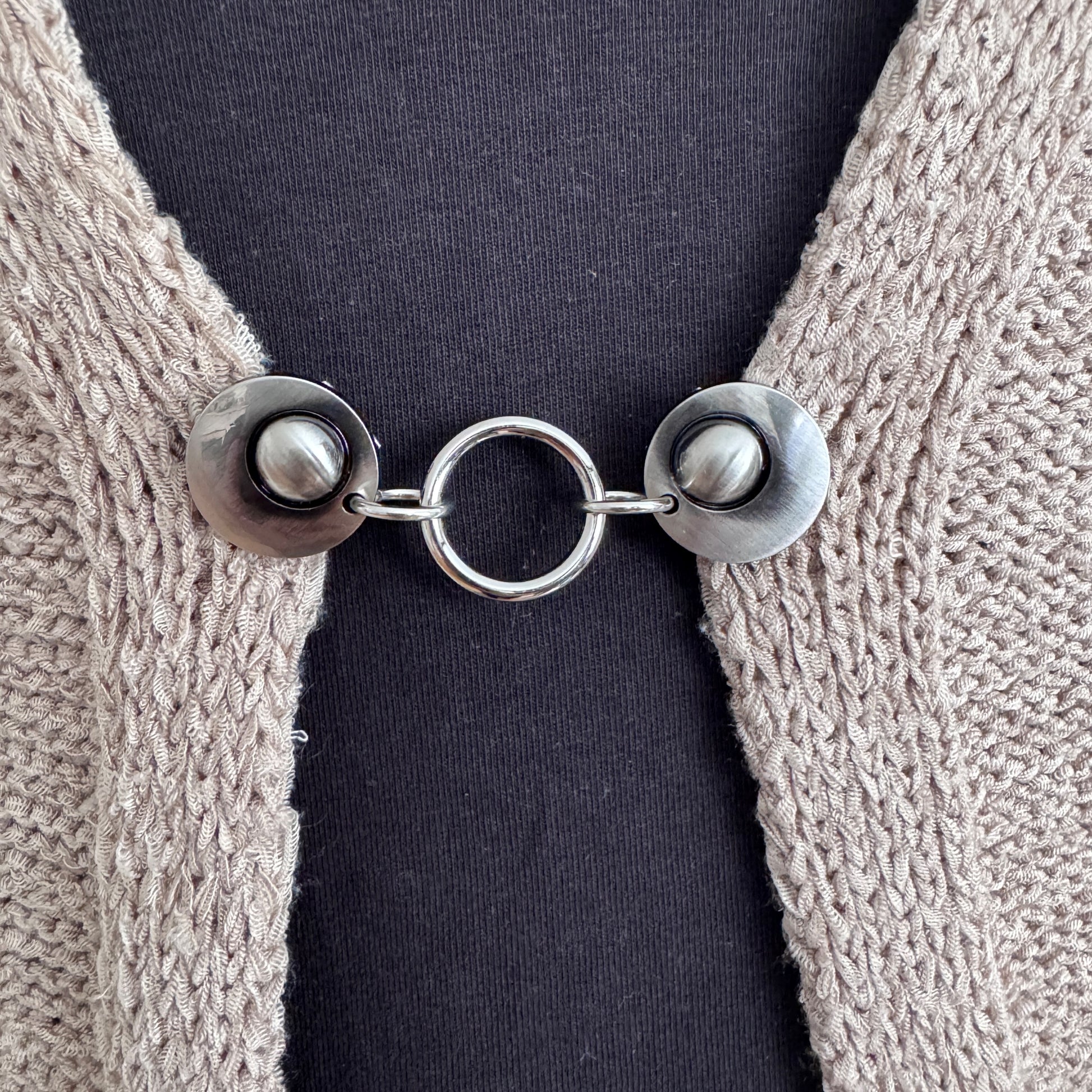 Vestsluiting Het Rondje zilver in close-up op een gebreid vest, elegante Fashionclip met zilverkleurige clips en ronde ringverbinding.