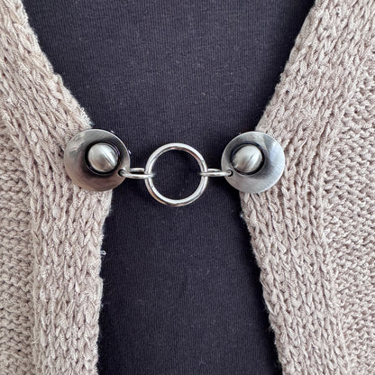Vestsluiting Het Rondje zilver in close-up op een gebreid vest, elegante Fashionclip met zilverkleurige clips en ronde ringverbinding.