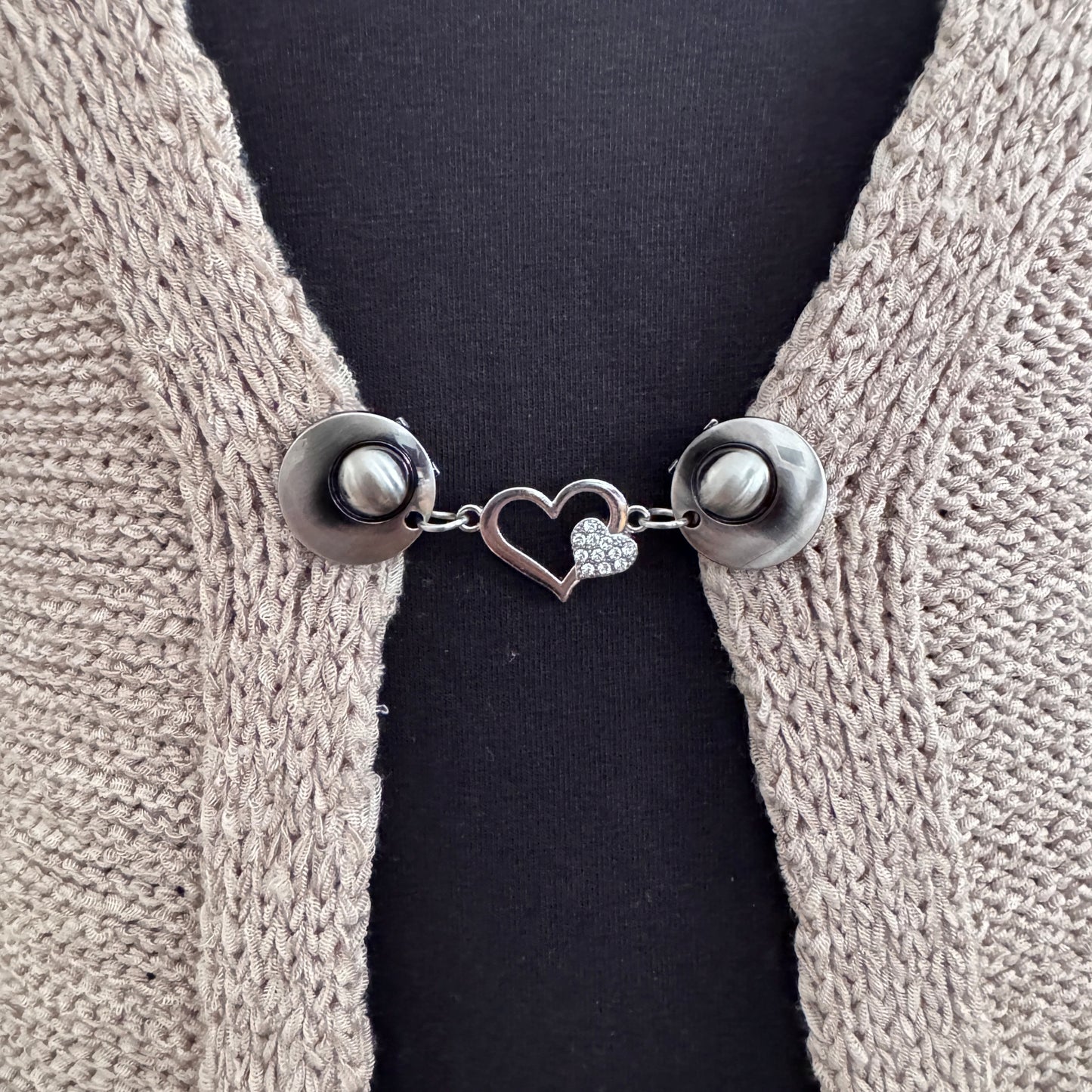 Vestsluiting Het Sprankelende Hart in close-up op een gebreid vest, elegante Fashionclip met zilverkleurige clips en hartvormige tussenbedel met strass.