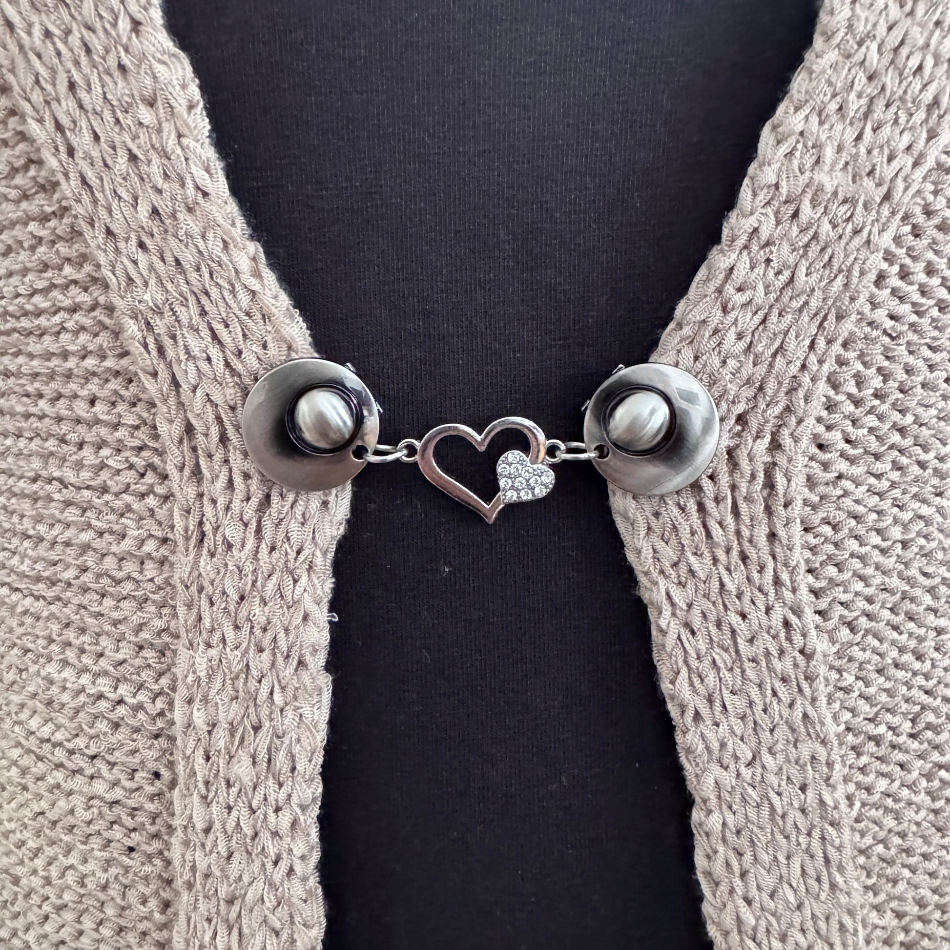 Vestsluiting Het Sprankelende Hart in close-up op een gebreid vest, elegante Fashionclip met zilverkleurige clips en hartvormige tussenbedel met strass.