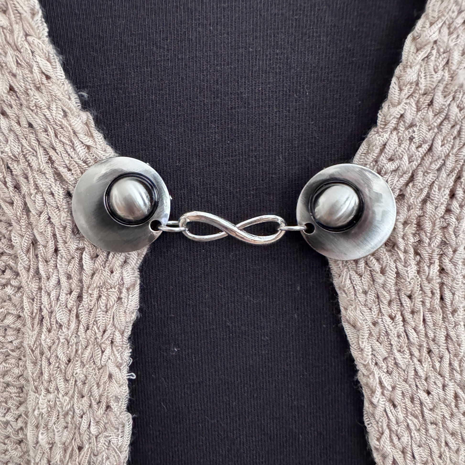 Vestsluiting Infinity zilver in close-up op een gebreid vest, elegante Fashionclip met zilverkleurige clips en infinity tussenbedel.