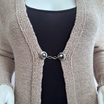 Vestsluiting Infinity zilver gedragen op een beige gebreid vest, elegante Fashionclip met zilverkleurige clips en infinity tussenbedel.