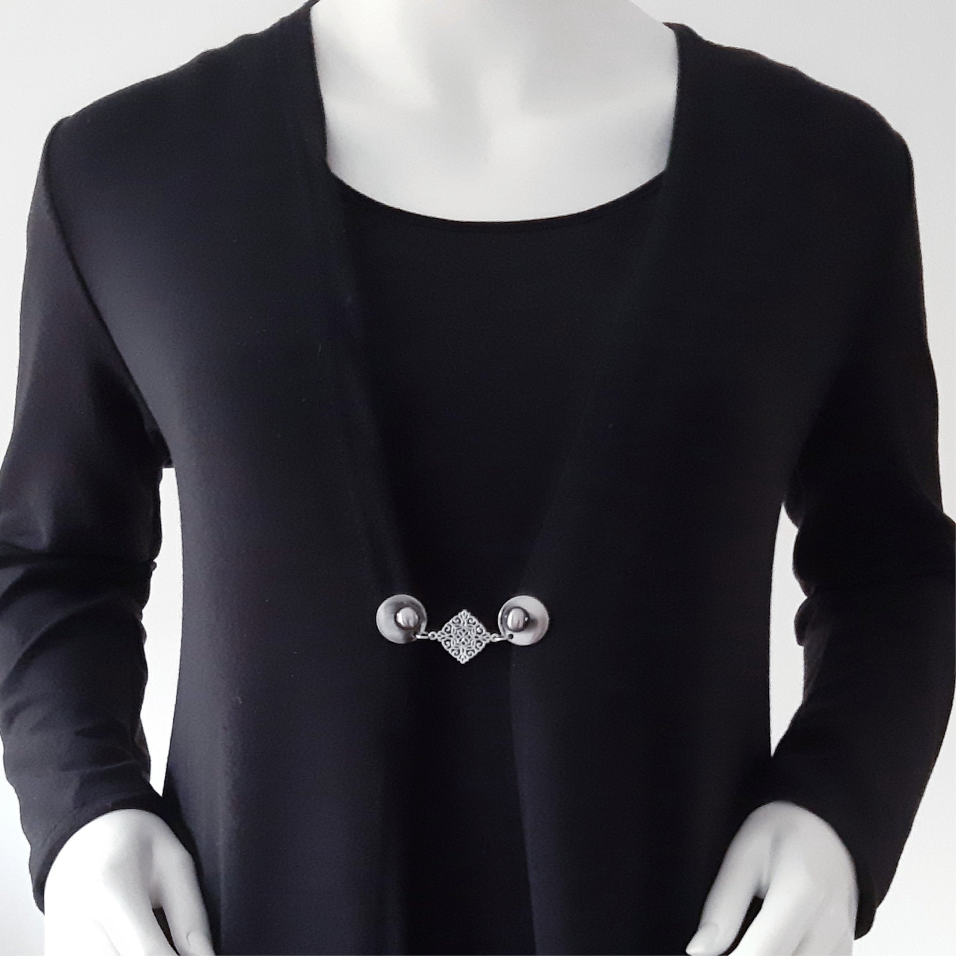 Vestsluiting met zilverkleurige clips en sierlijke ruitvormige tussenbedel, elegante Fashionclip gedragen op een zwart vest.