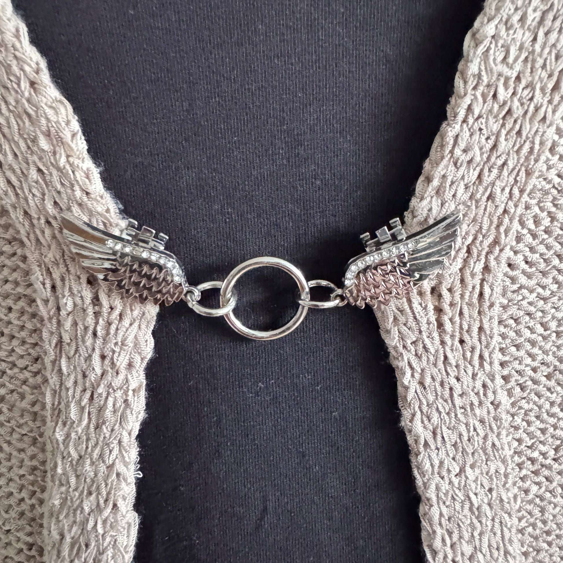 Vestsluiting Vleugels van Vrijheid zilver in close-up op een gebreid vest, elegante Fashionclip met vleugelvormige clips en ronde ringverbinding.